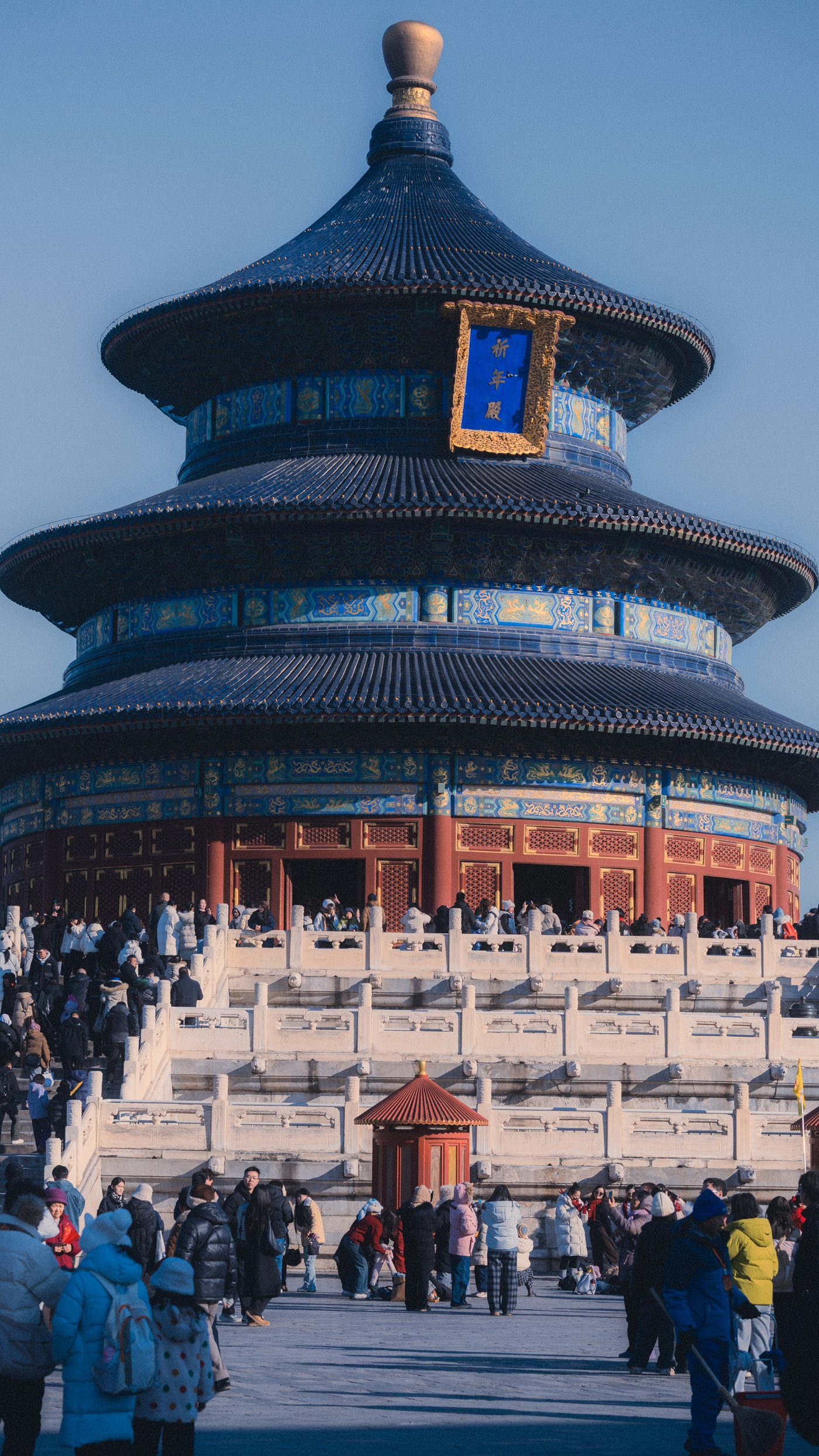 Crowds in winter coats fill the terraces below a round triple eaved temple hall, its blue tiled roofs lit by low sun.
