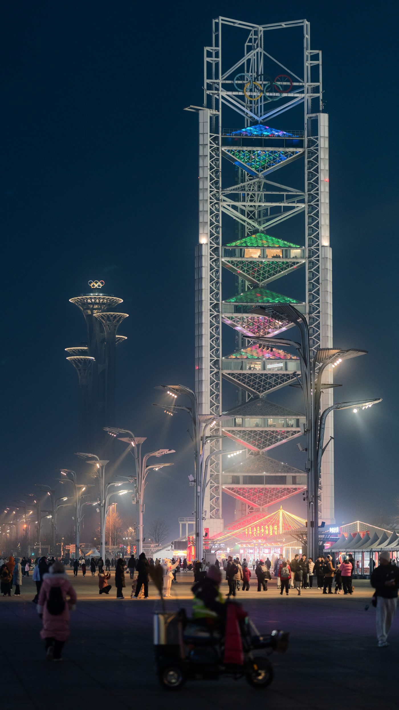 Lattice observation tower crowned with five interlocking rings glows green and pink above a night plaza where people cross in padded coats.