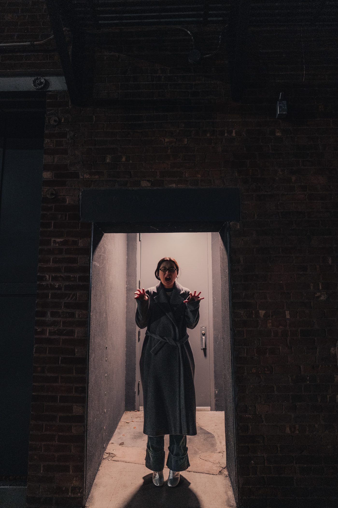 Woman in a long grey belted coat stands in a lit doorway of a dark brick wall, mouth open wide, hands raised and clawed.