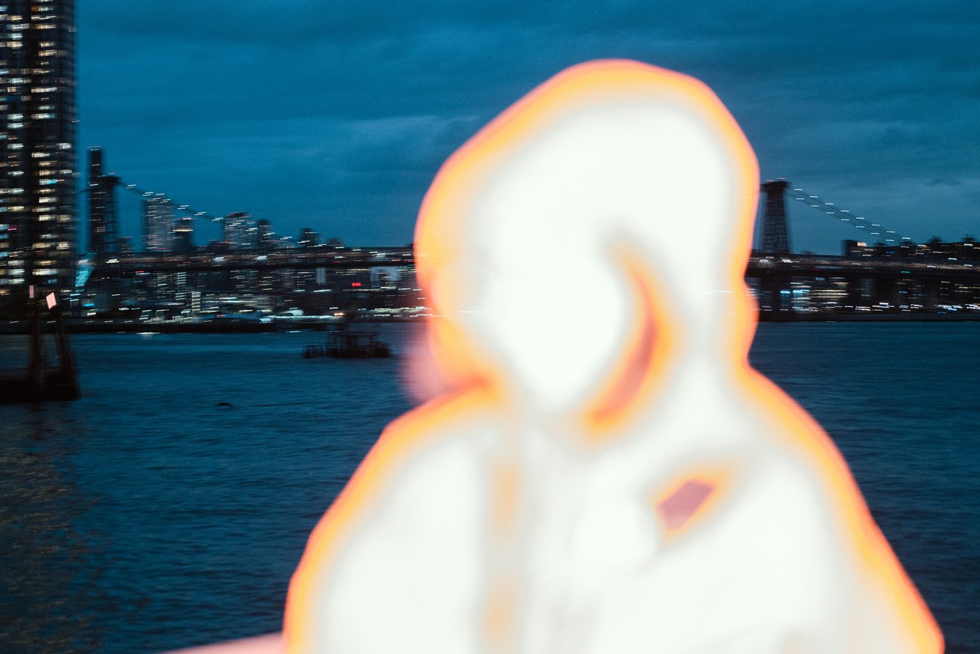 A hooded figure blown out to pure white and far out of focus fills the foreground, a lit suspension bridge and dusk river behind.