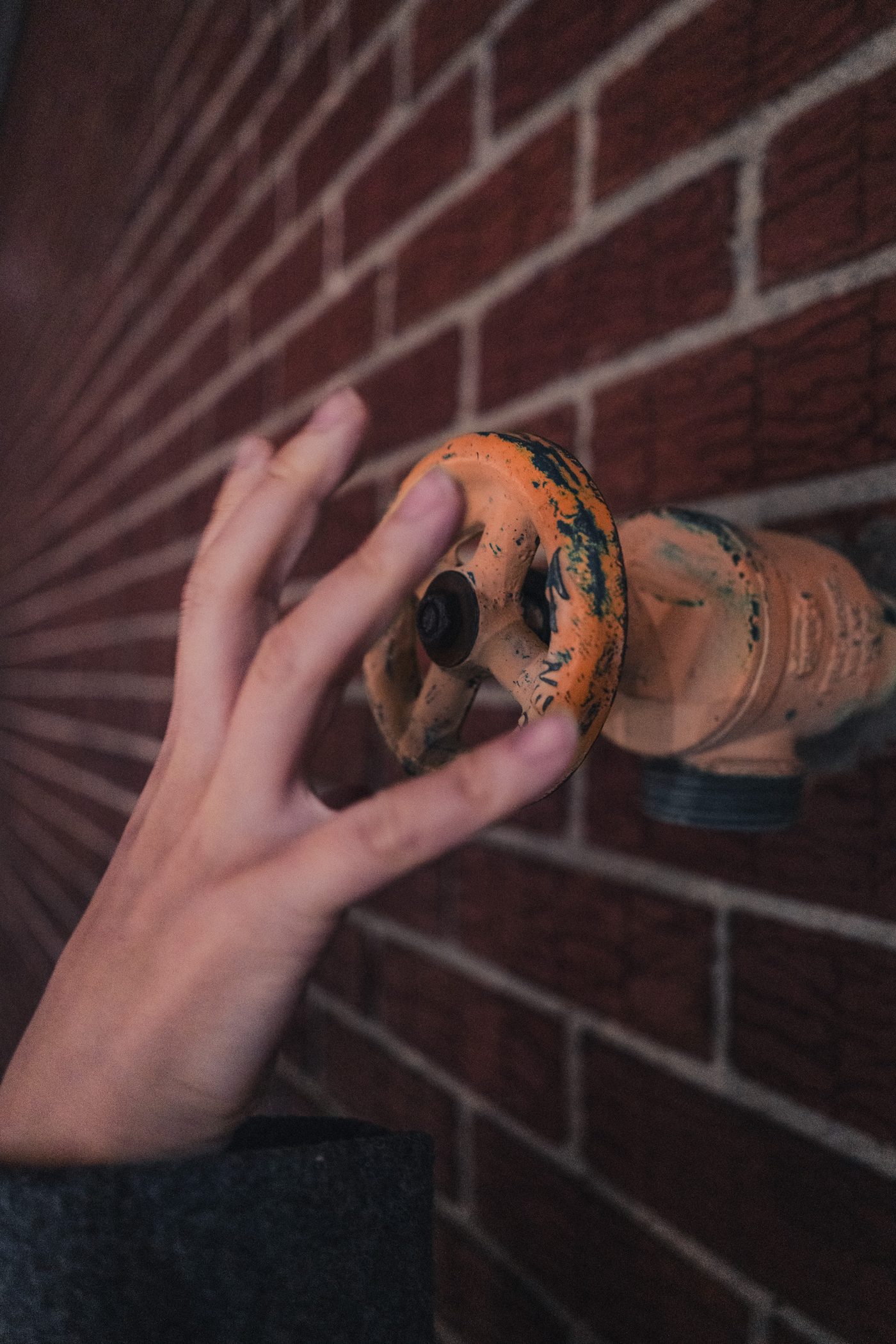 A hand with fingers spread reaches toward a chipped orange standpipe valve set into red brick, the wall receding into dim evening light.
