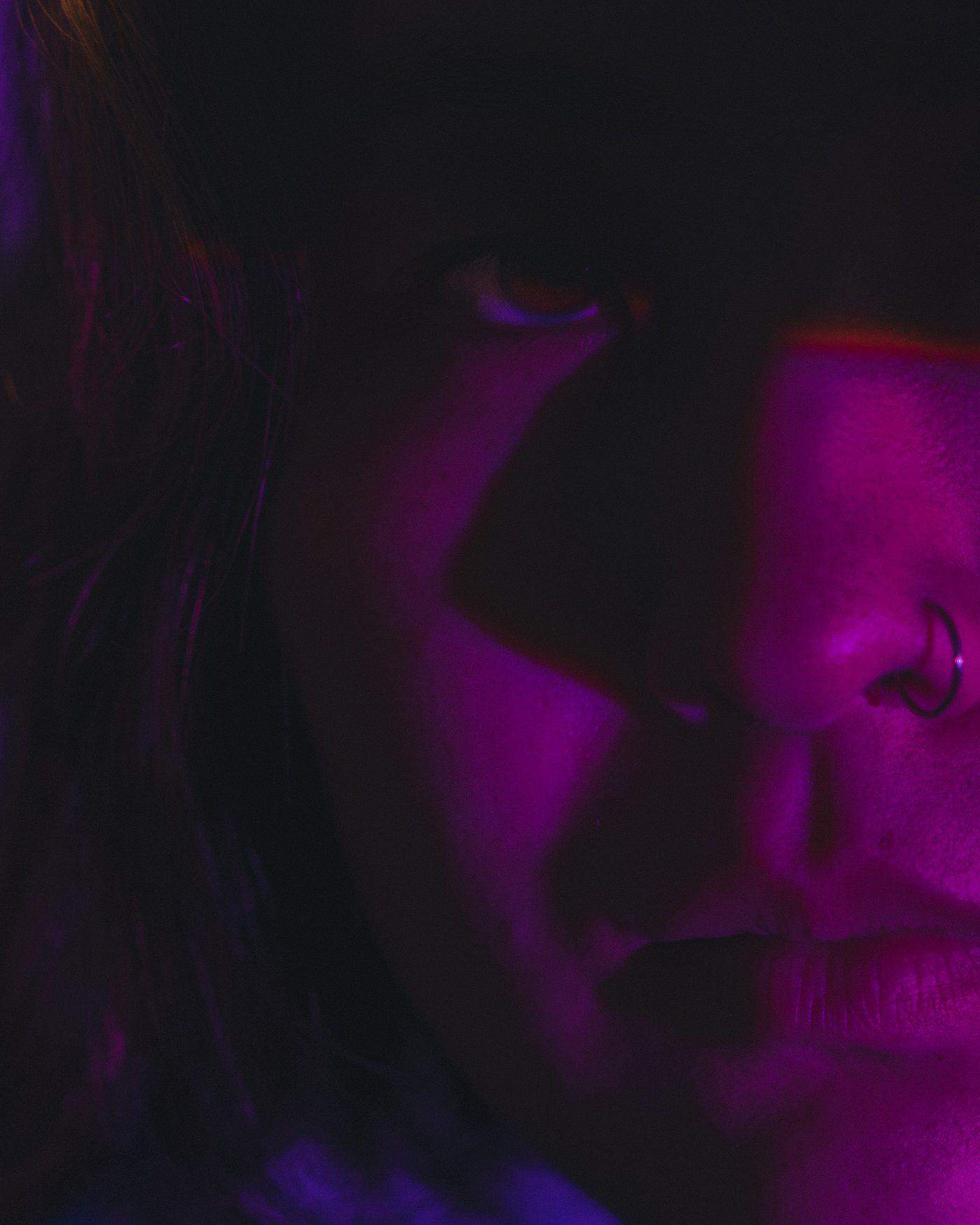 Close profile of a woman's face lit in magenta, one eye sunk in shadow, a metal hoop through her nostril.
