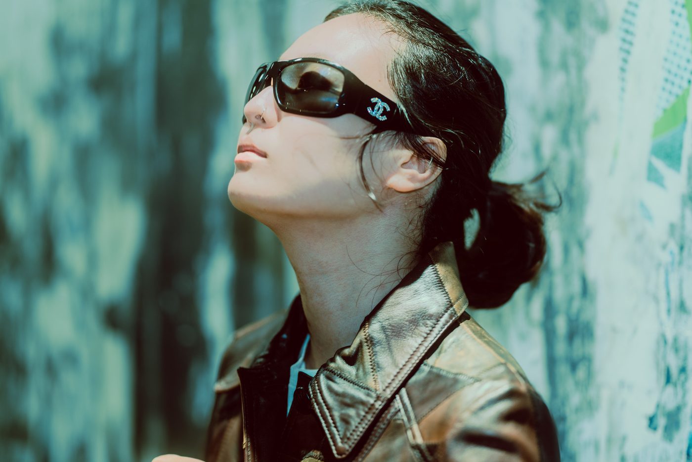 A woman tilts her chin up in profile behind dark wraparound sunglasses, daylight raking her throat, weathered painted wall behind.