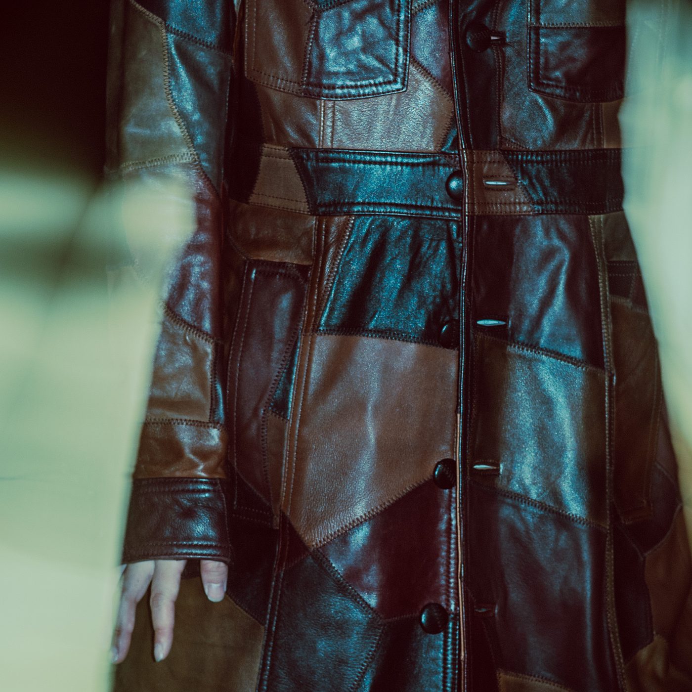 Patchwork leather coat in brown and black panels, zigzag stitching and dark buttons running down the front, a pale hand at the cuff.
