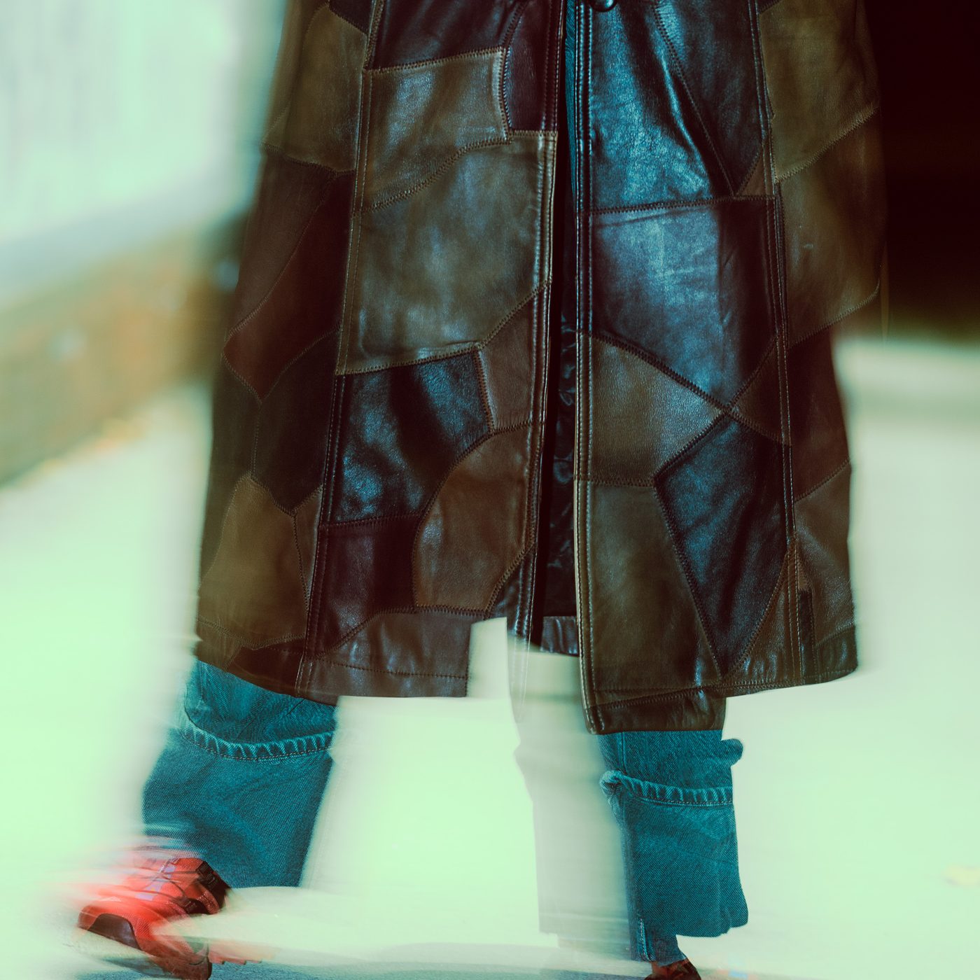 Leather coat hem swings mid-stride over cuffed blue jeans, one red sneaker smeared into motion blur against pale ground.
