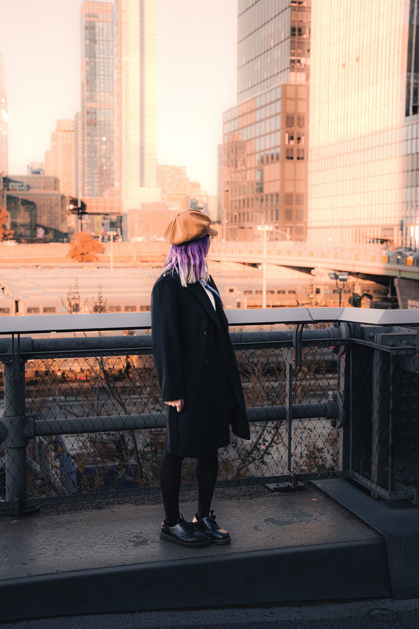 A woman in a long black coat and tan newsboy cap stands in profile at a railing, low sun washing the towers behind her.