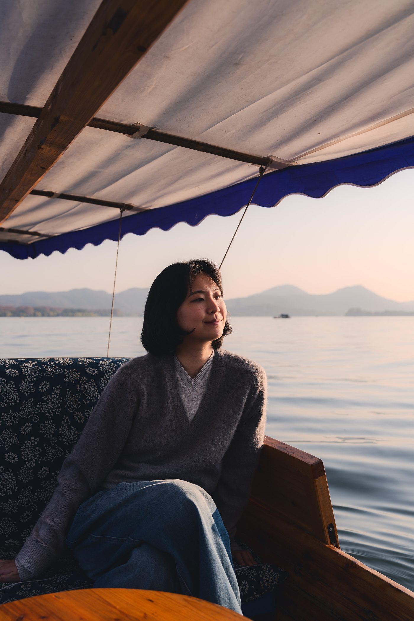 A woman in a grey sweater sits under a boat's blue-trimmed canopy, half smiling as she looks across still water to hazy hills.