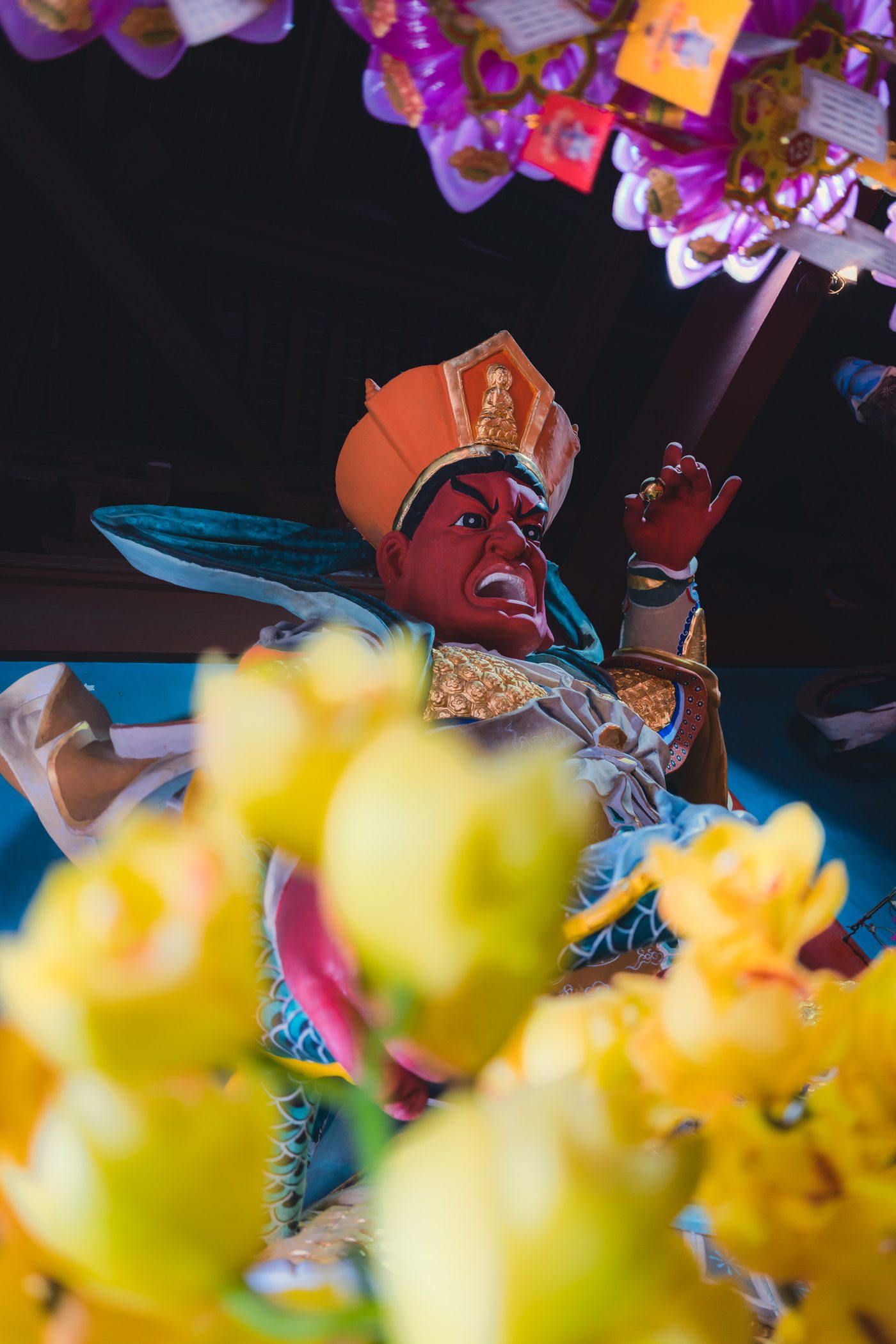 Red-faced temple guardian statue snarls with one hand raised, pink lotus lamps overhead and yellow offering flowers blurred across the foreground.