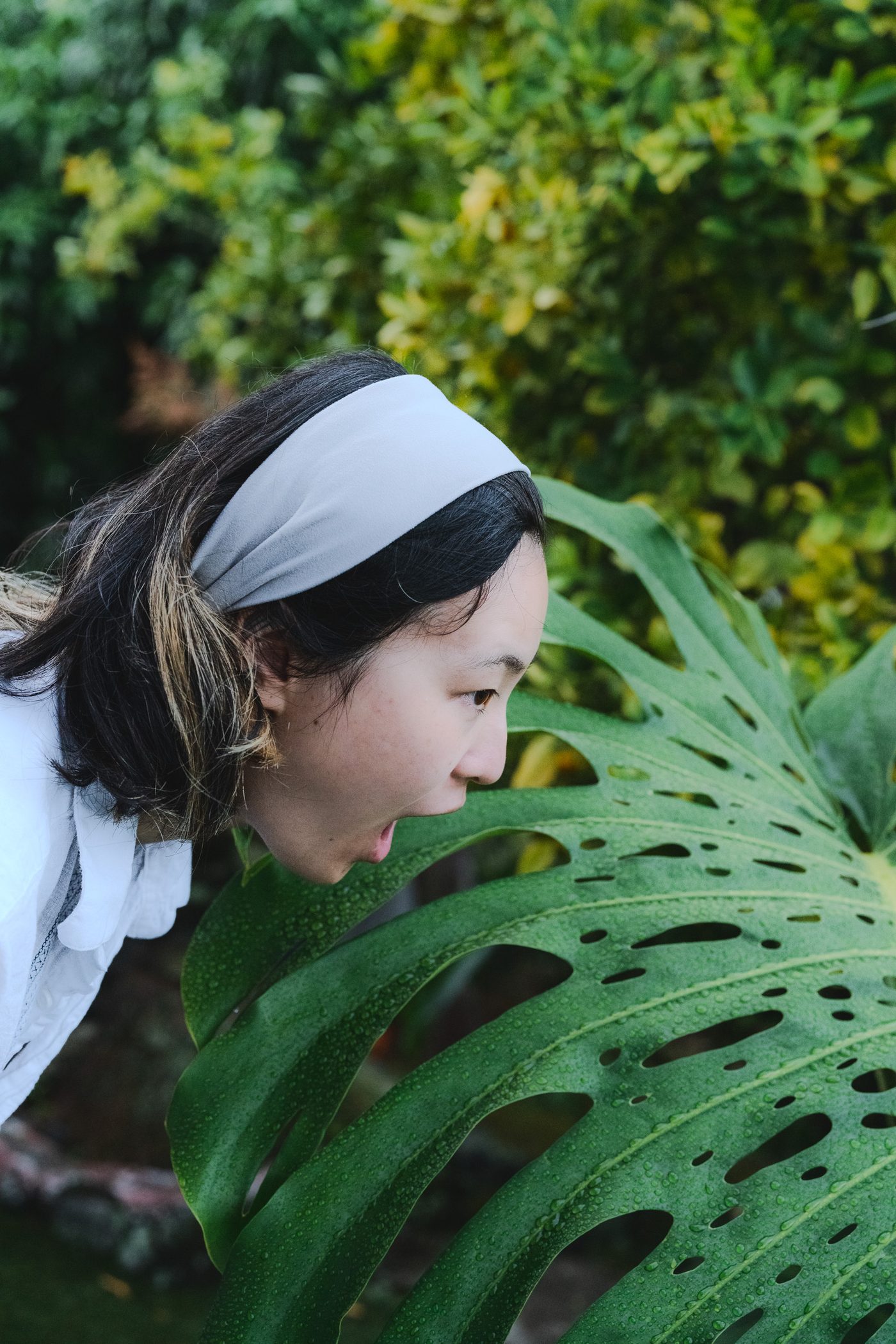 A woman with bleached streaks in her hair leans down open-mouthed to a rain-beaded monstera leaf, soft overcast light on her profile.