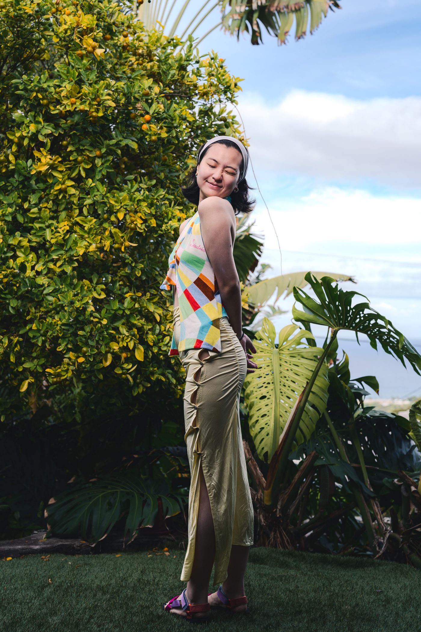 DQ turns to look back over her shoulder in a patchwork halter top and long sage skirt, a fruiting citrus tree crowding the frame.