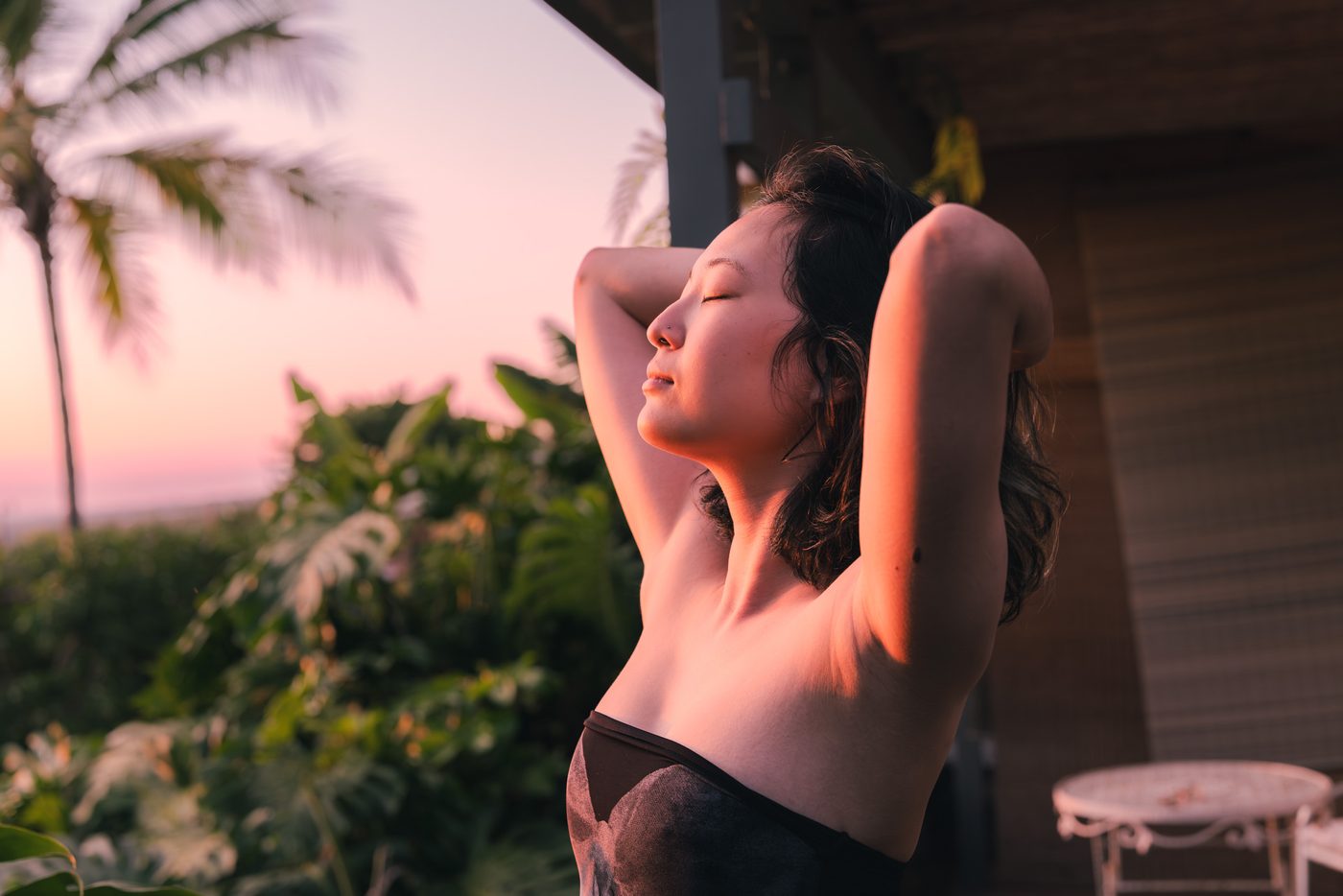 A woman stands with both arms raised behind her head and eyes closed, warm pink sunset light on her shoulders, palm fronds blurred behind.