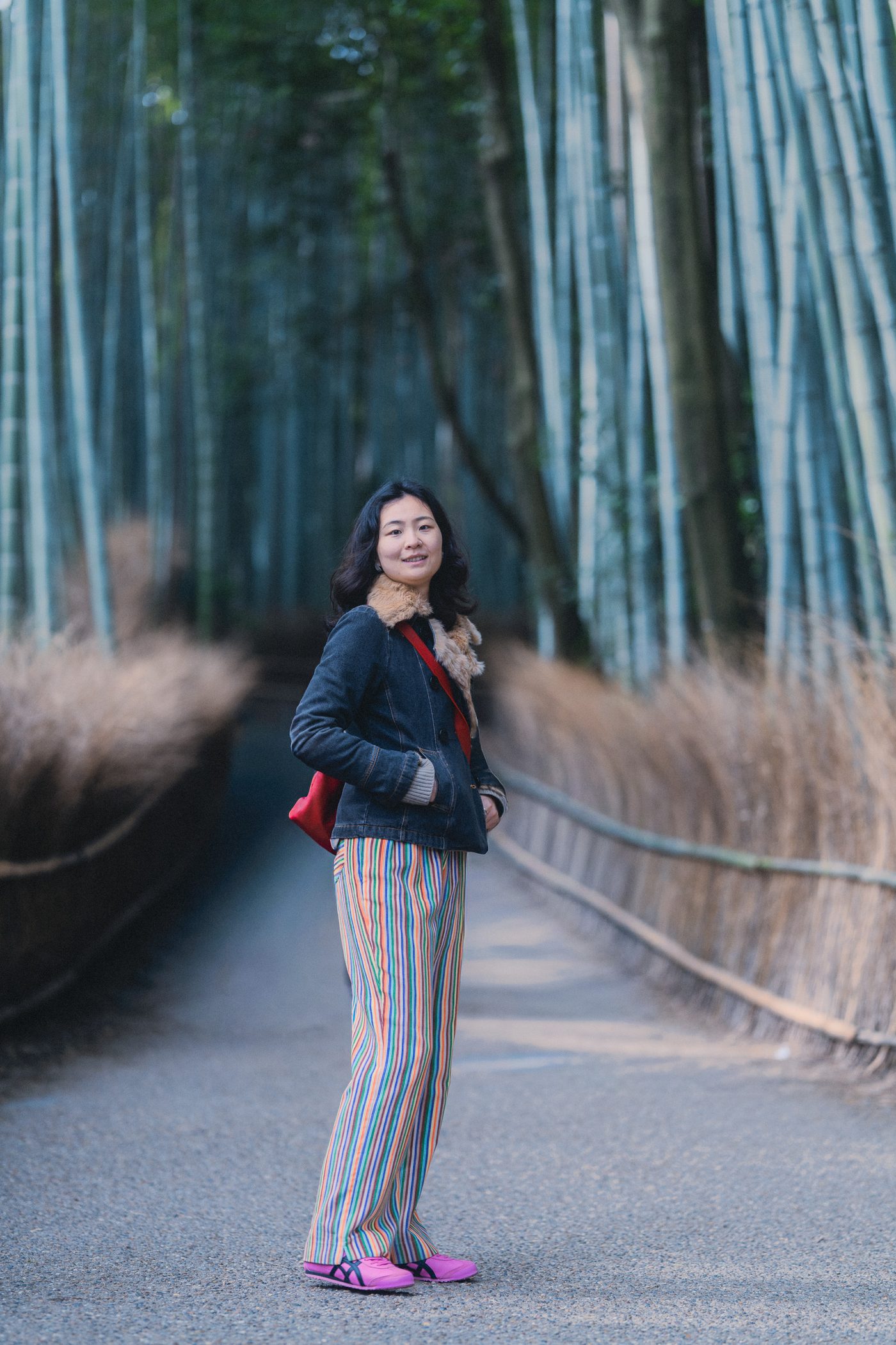 A woman in a denim jacket and rainbow striped trousers pauses on a paved path through tall bamboo, turning back into soft shaded light.