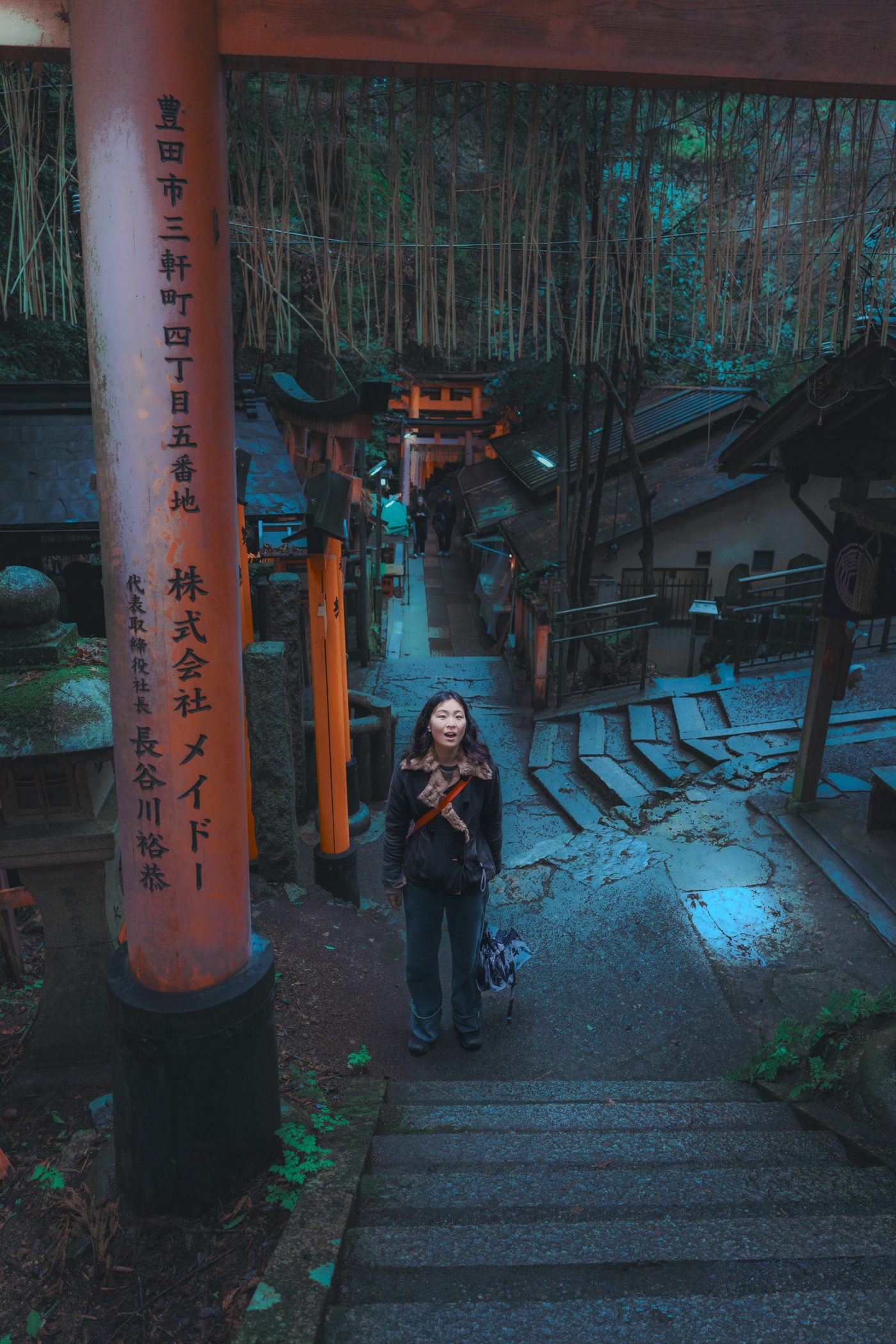 A woman in a dark shearling coat stands open mouthed at the top of wet stone steps, a vermilion torii gate rising over her at dusk.