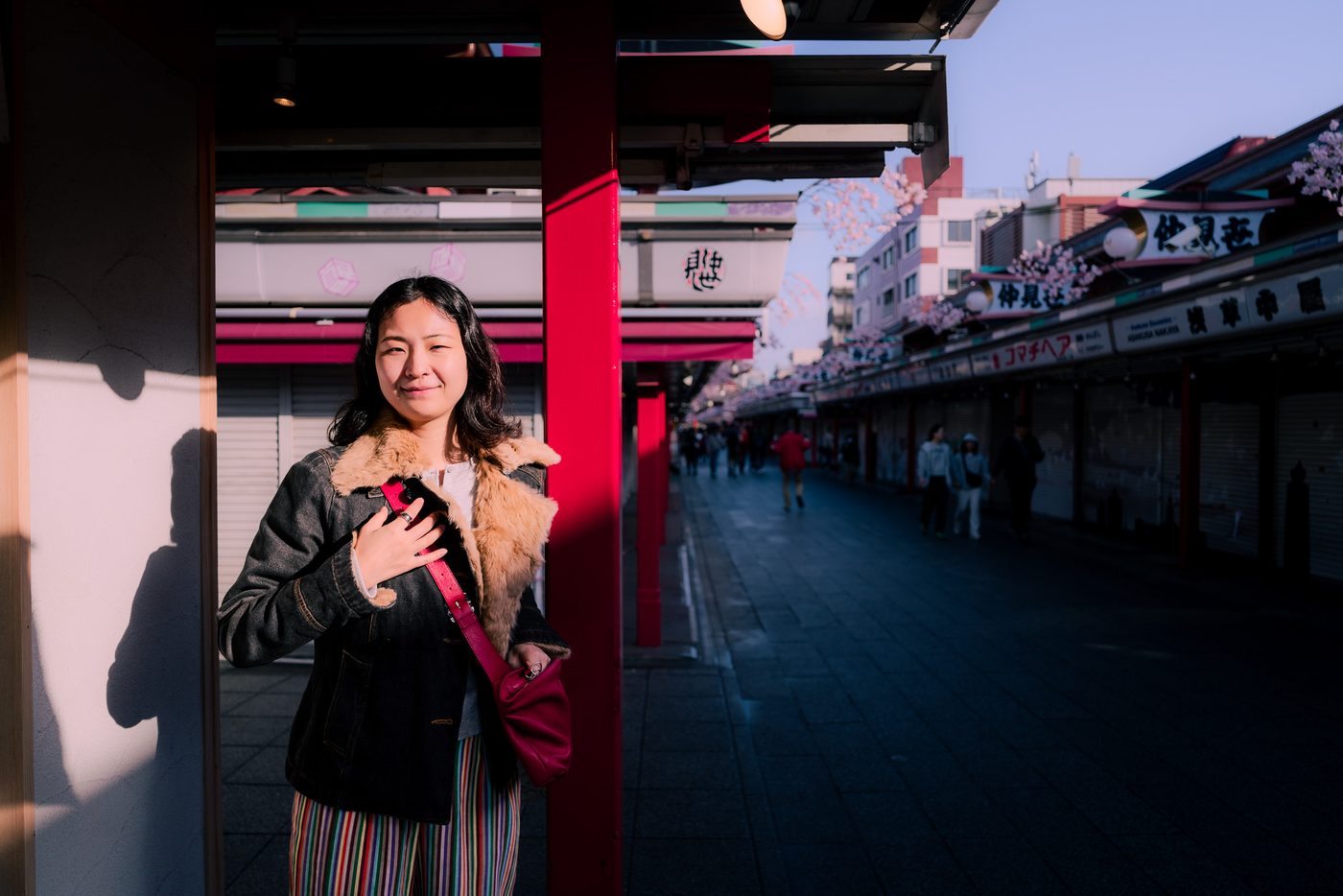 A woman beside a red pillar on a shuttered market street holds her magenta bag strap, low late sun raking her face and the wall behind.