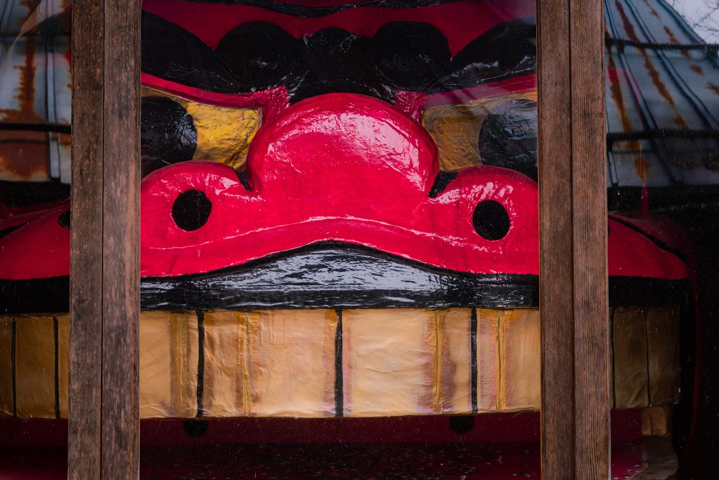 Glossy scarlet and gold ornament sits inside a display case behind weathered wooden bars, two black eye holes above a pleated gold panel.
