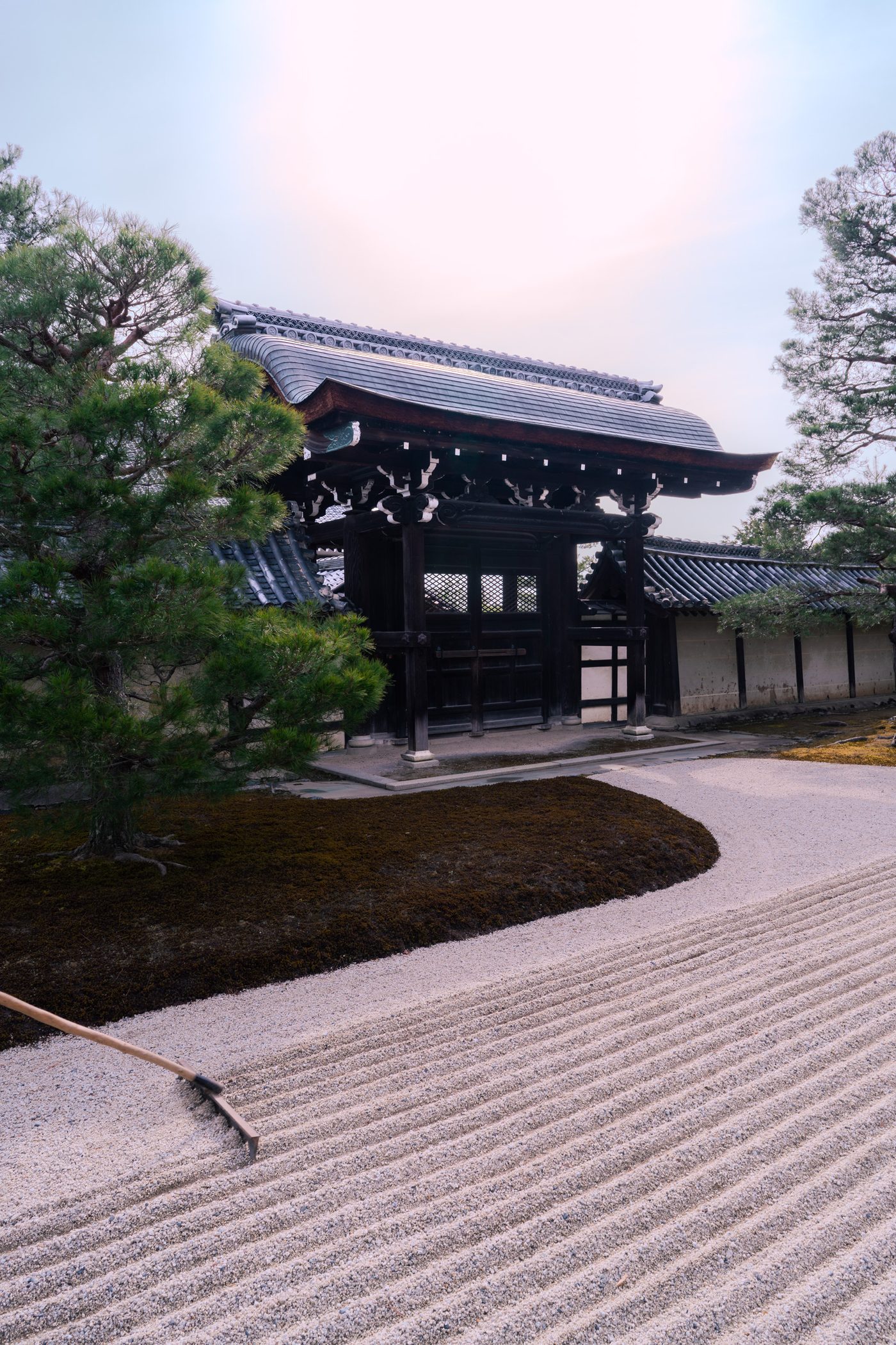 A dark timber temple gate stands behind raked white gravel, its curved tile roof lit by flat hazy sun, a rake left in the foreground.