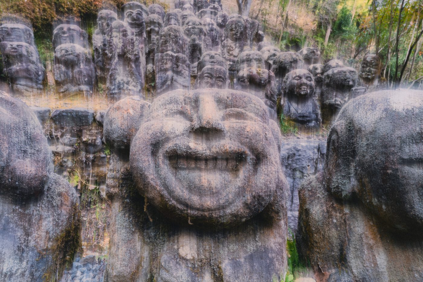 Rows of moss-crusted stone figures crowd a hillside, one broad grinning face filling the foreground while vertical streaks blur the ranks behind.