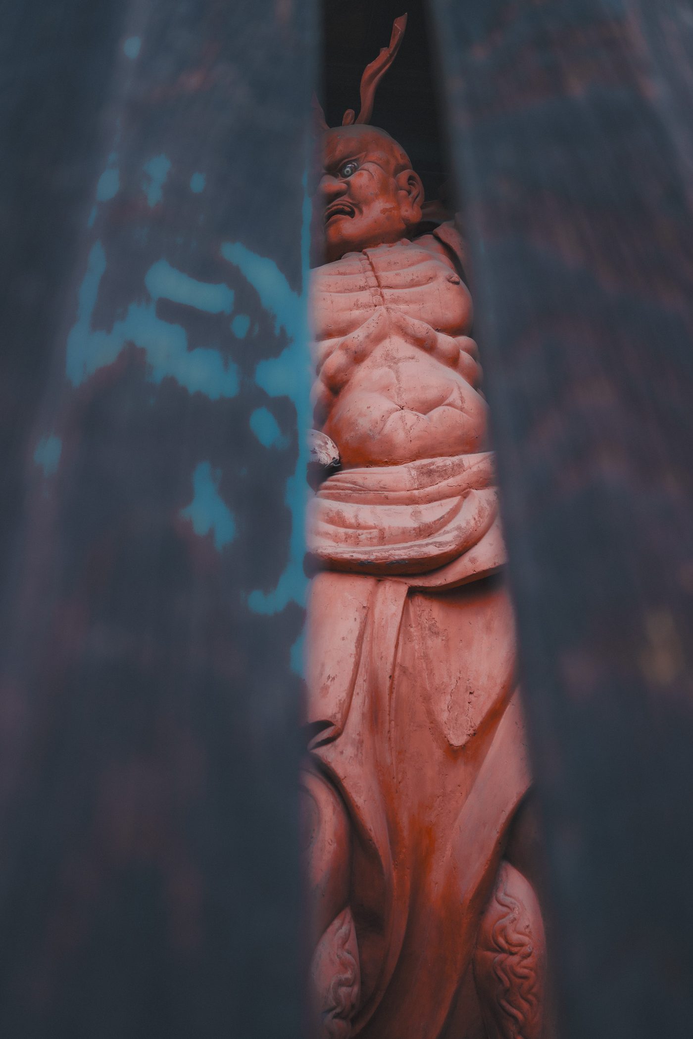 A red-painted temple guardian statue scowls through a narrow vertical gap between dark timber pillars, muscled torso caught in a slot of light.