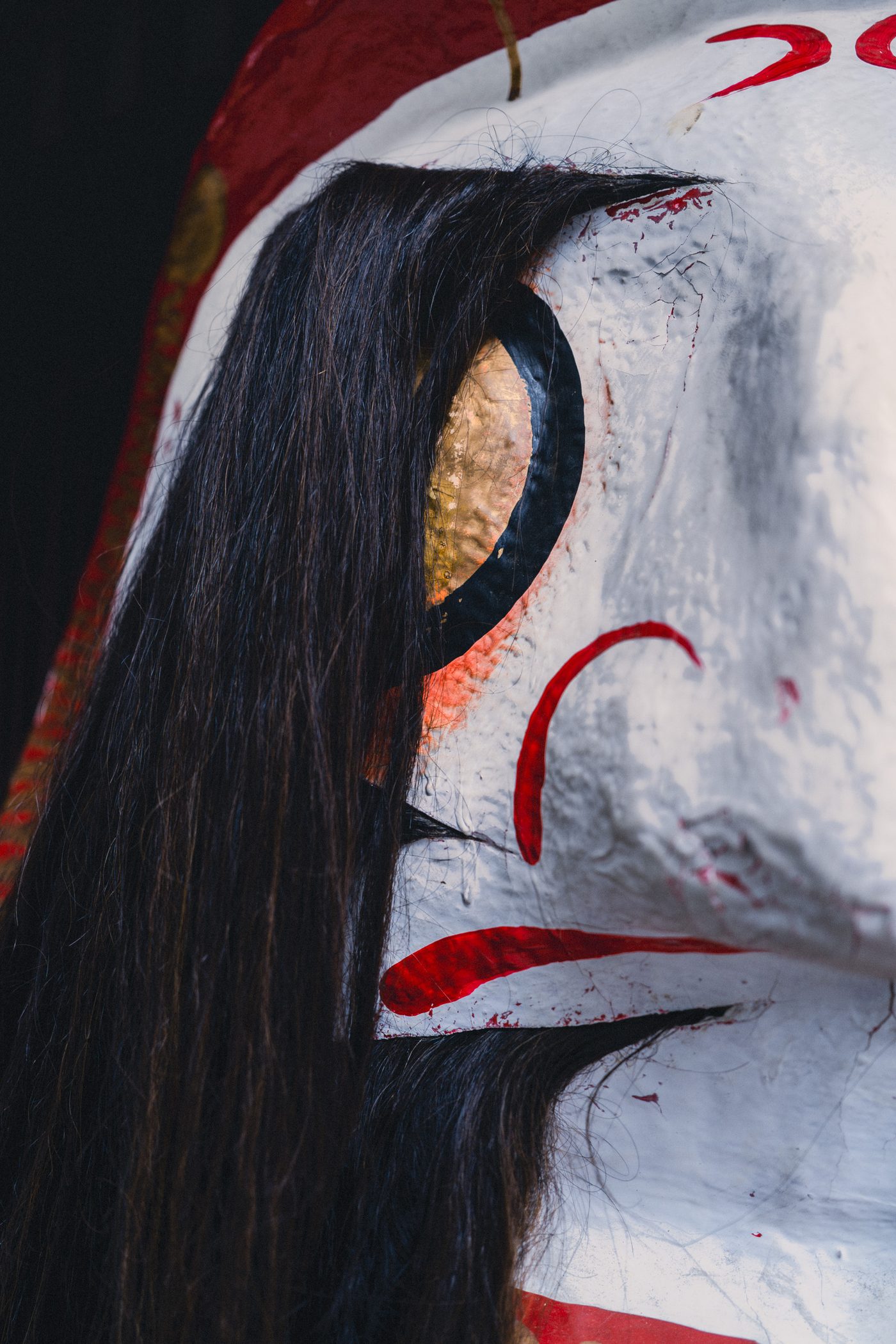 A white-lacquered festival mask fills the frame, strands of real black hair falling over a gold leaf eye and red brow strokes.