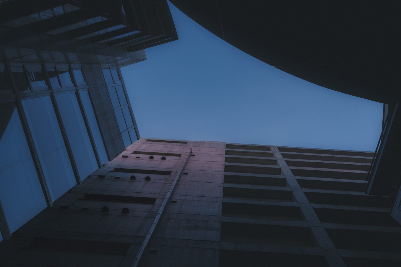 Bare concrete walls converge overhead as a drainpipe climbs toward a wedge of cold blue evening sky, the whole frame sunk in shadow.