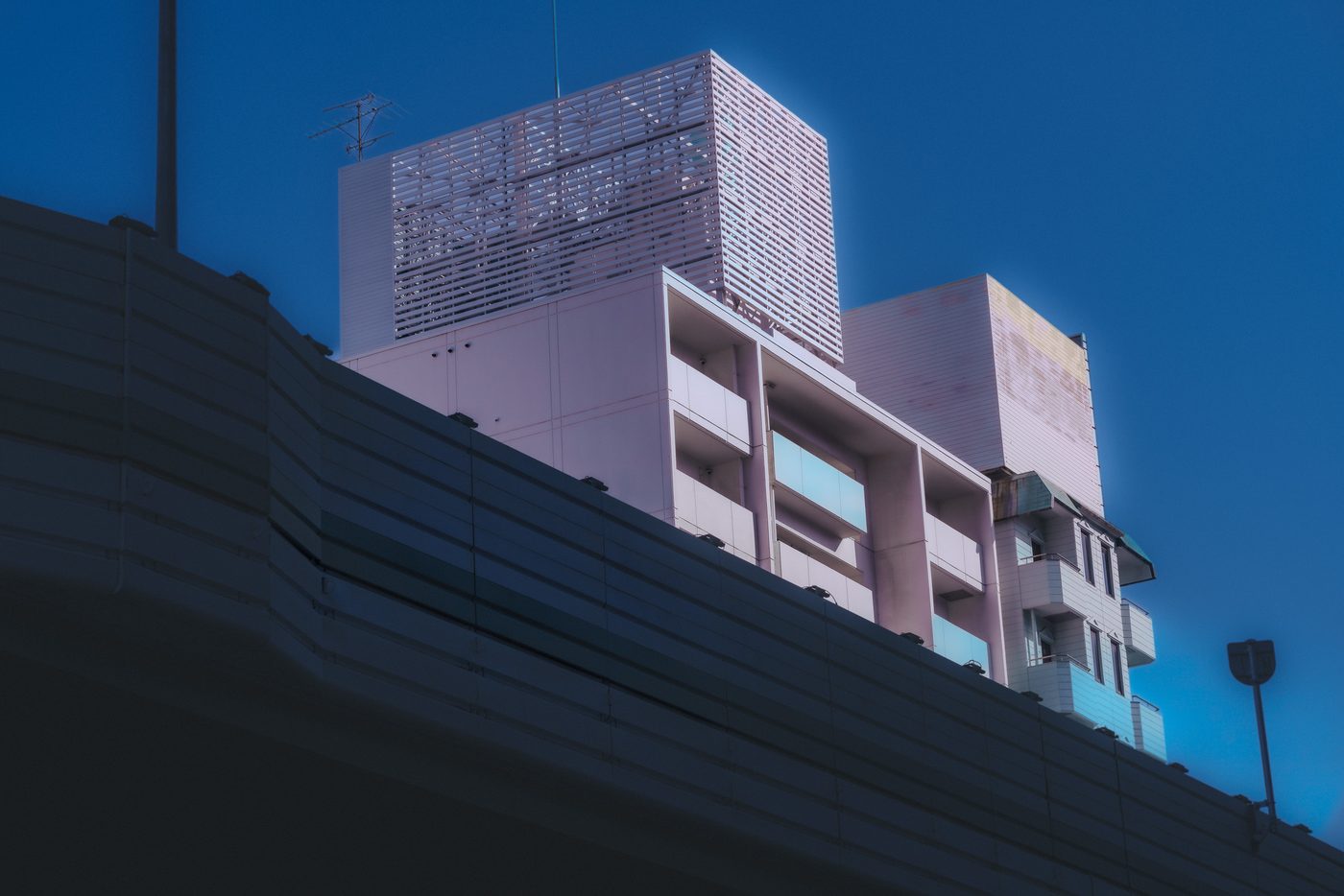Pale pink apartment blocks and a slatted rooftop screen rise over the dark curve of an elevated roadway wall against deep blue dusk.