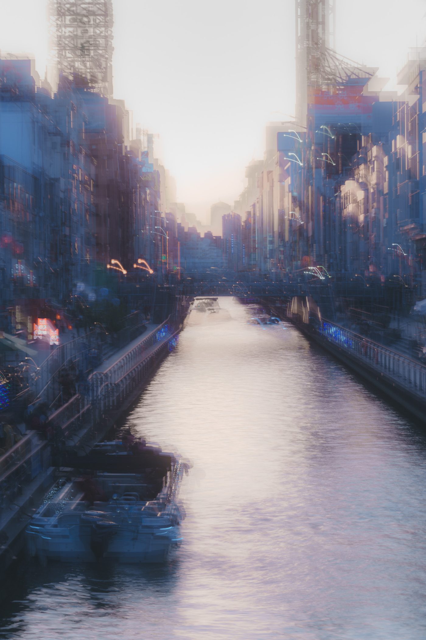 A canal runs straight between signboard-covered buildings toward a white blown-out sky, moored boats and street lights doubled into soft overlapping smears.