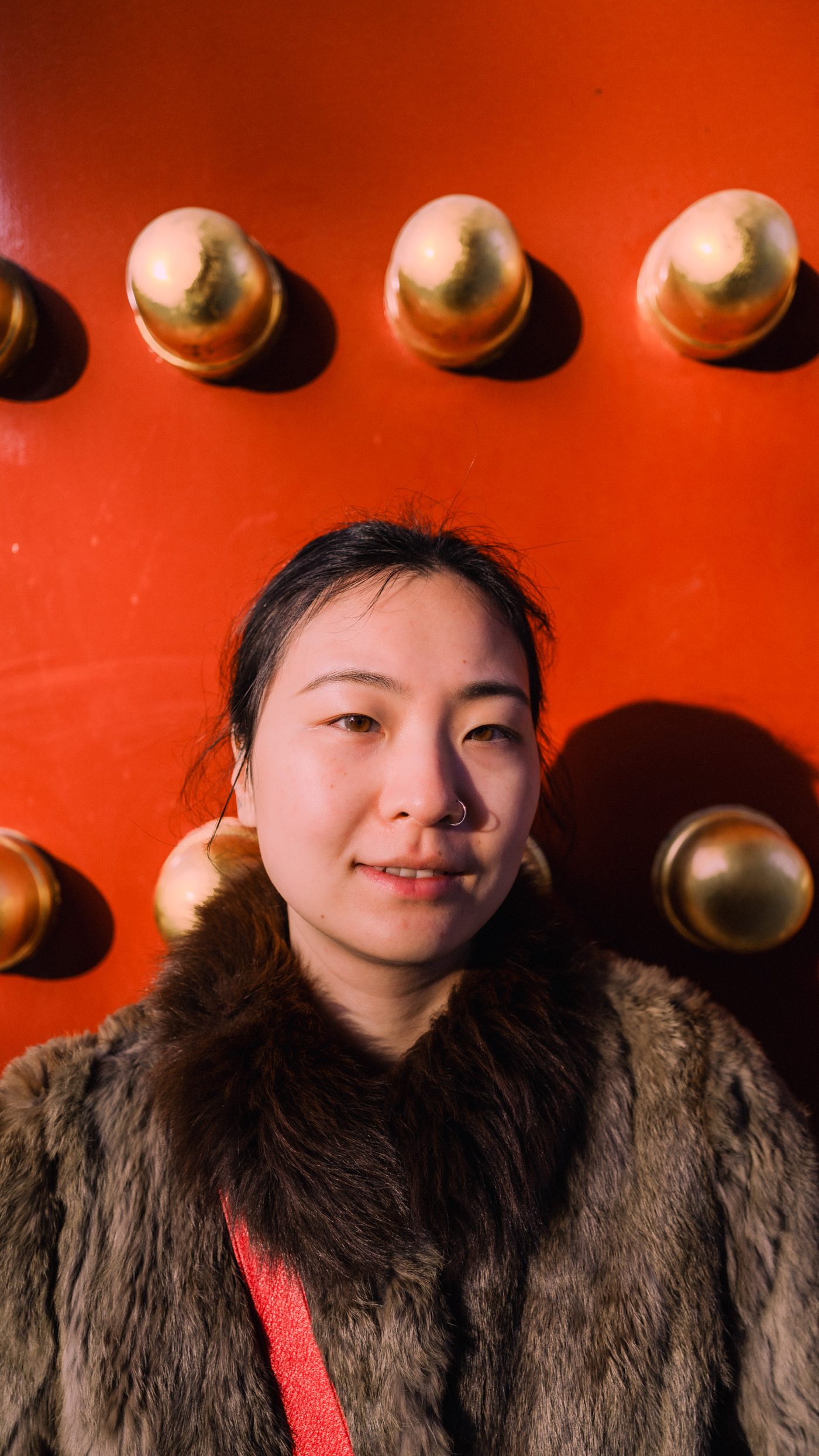 A woman in a grey fur coat smiles faintly against a vermilion temple door, hard low sun raking the gold bosses above her.