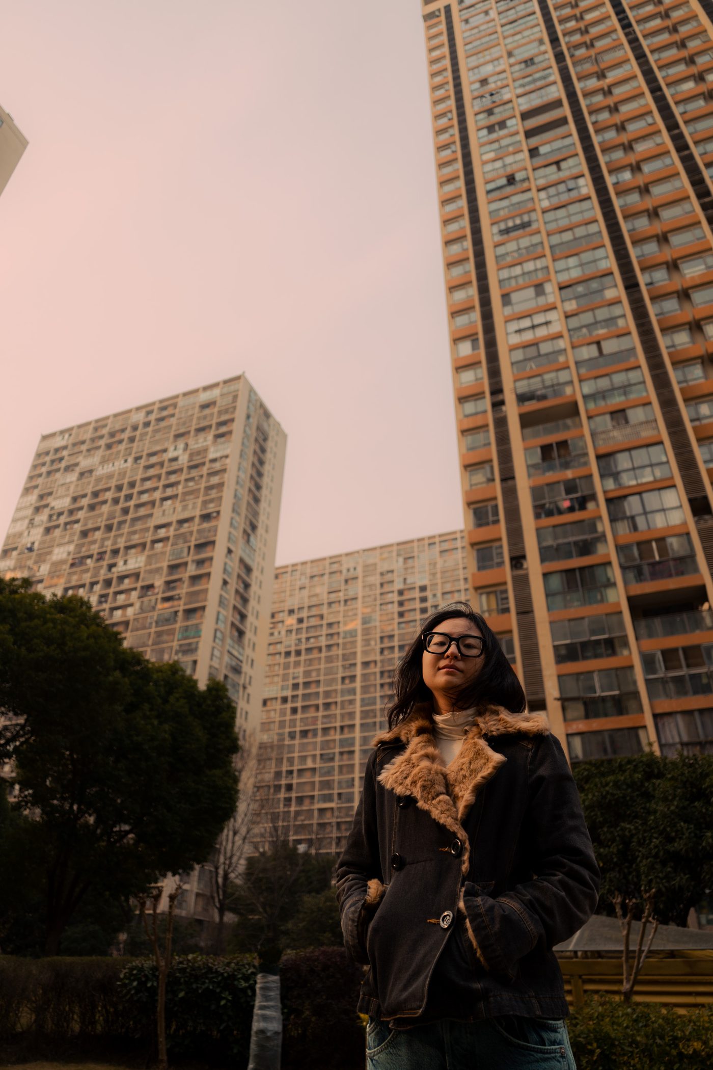 Bespectacled woman in a shearling collared denim coat stands beneath three concrete apartment towers, hands pocketed, sky a hazy pink.