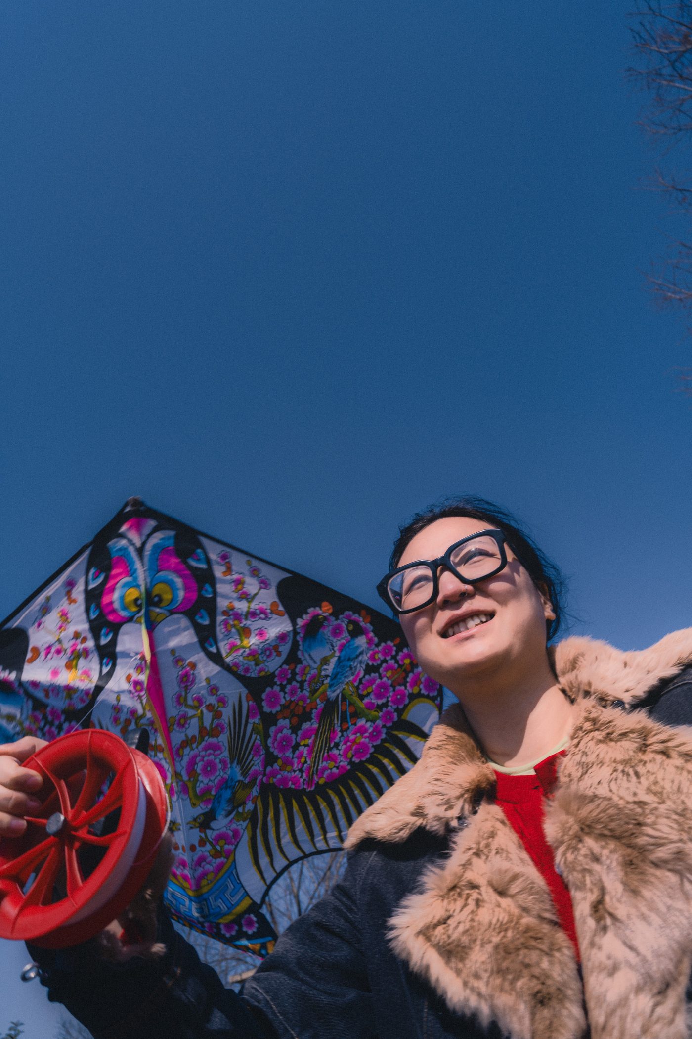 Grinning woman in a fur collar coat holds a red kite reel, a printed butterfly kite lifting beside her into deep blue sky.