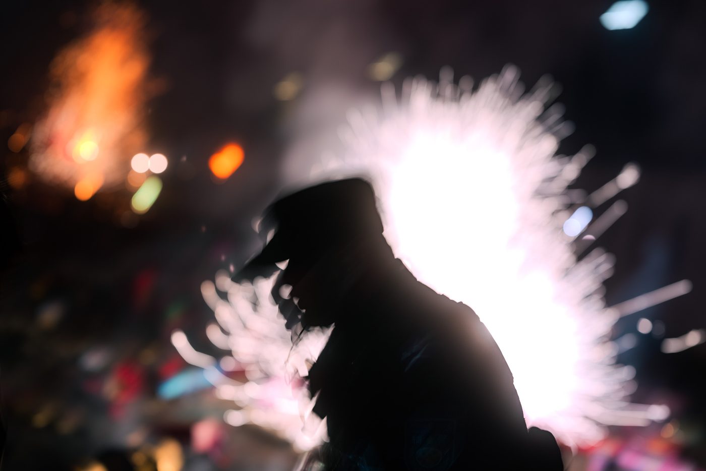 Silhouetted figure in a peaked cap set against a burst of white firework sparks, blurred orange street lights scattered through the dark.