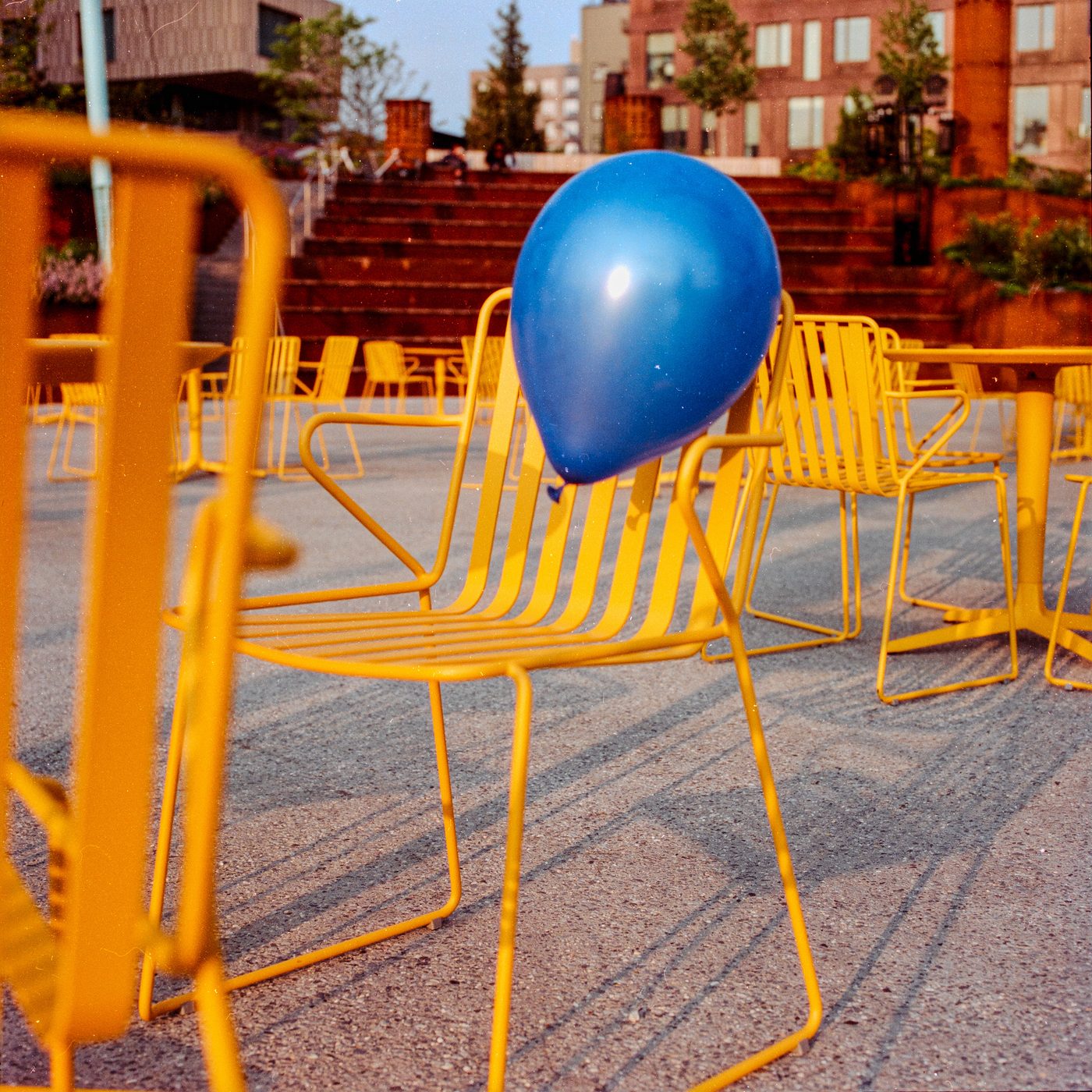 Blue balloon resting on a yellow wire cafe chair, low sun raking across gravel and empty tables on a plaza.