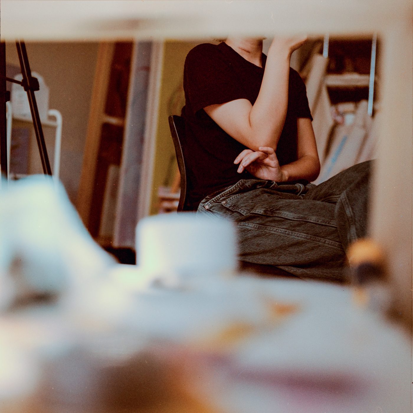 Woman in a black tee sits with arms crossed over her knees, head cropped off, seen low across a blurred tabletop.