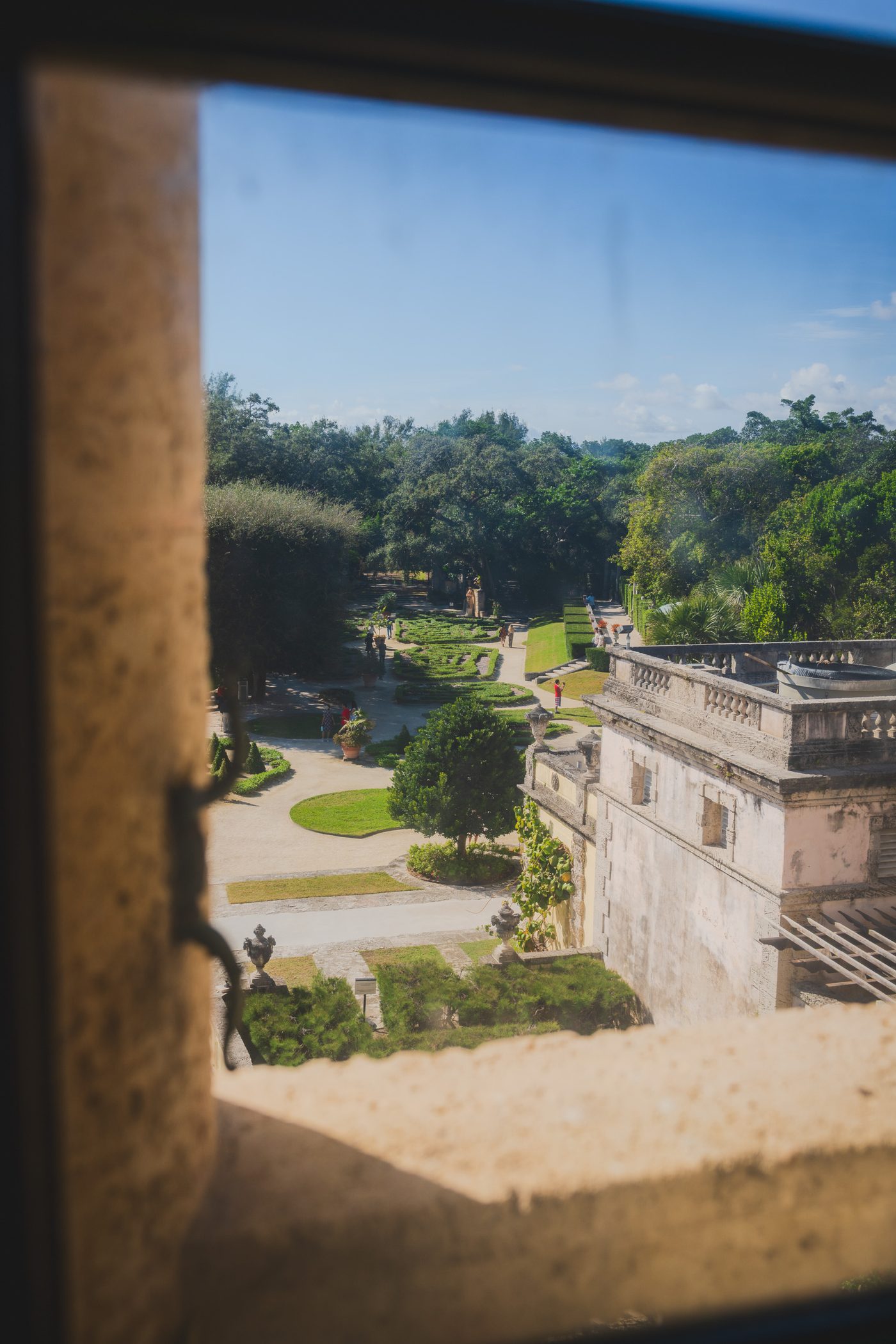 Clipped garden parterres and a weathered stone terrace seen through a blurred window opening, bright midday sun on the gravel paths below.