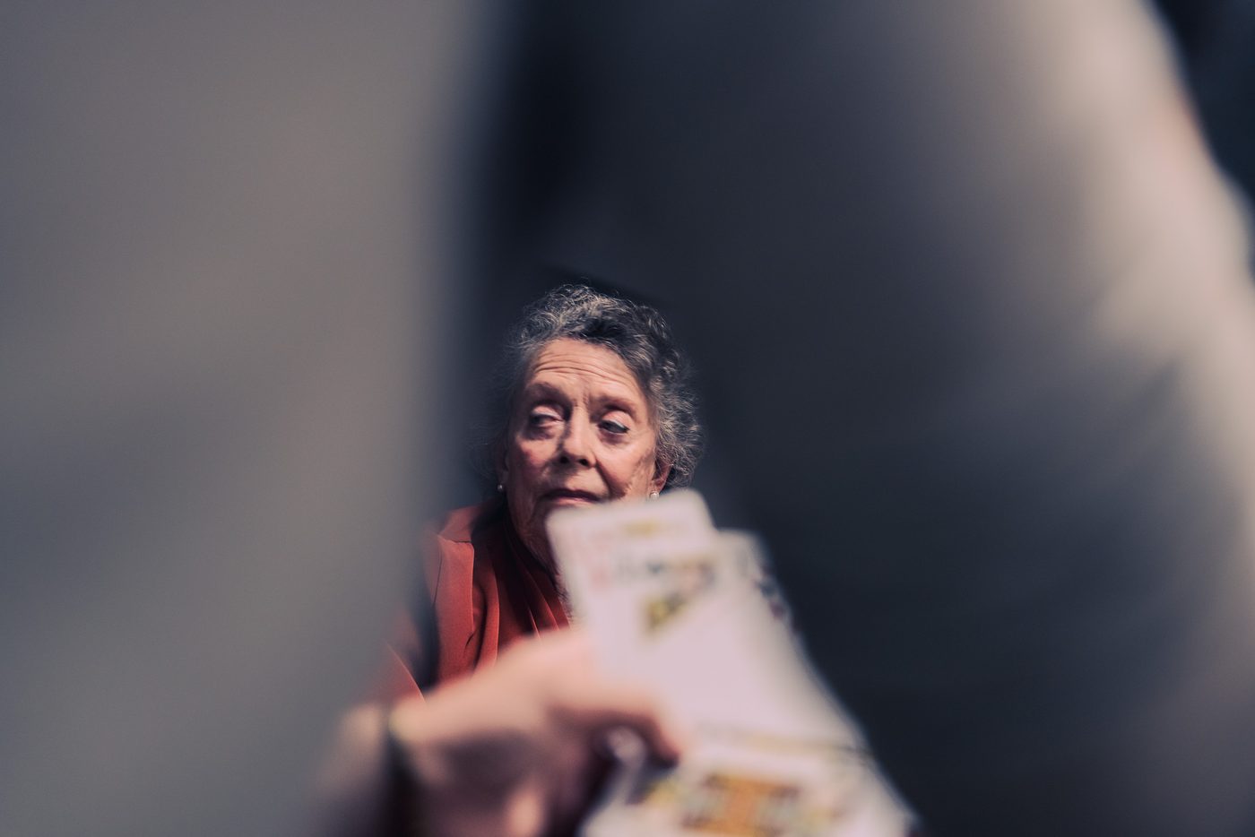 An older woman in a rust blazer glances sideways through a gap between dark blurred shapes, a hand of playing cards raised in front.