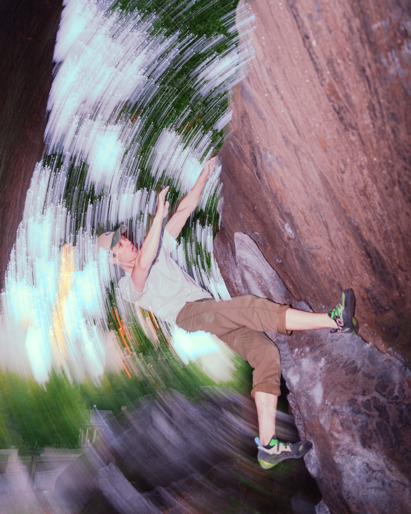 A climber in a cap and white tee stretches for a high hold on red brown rock, the leafy background swirled into radial streaks.