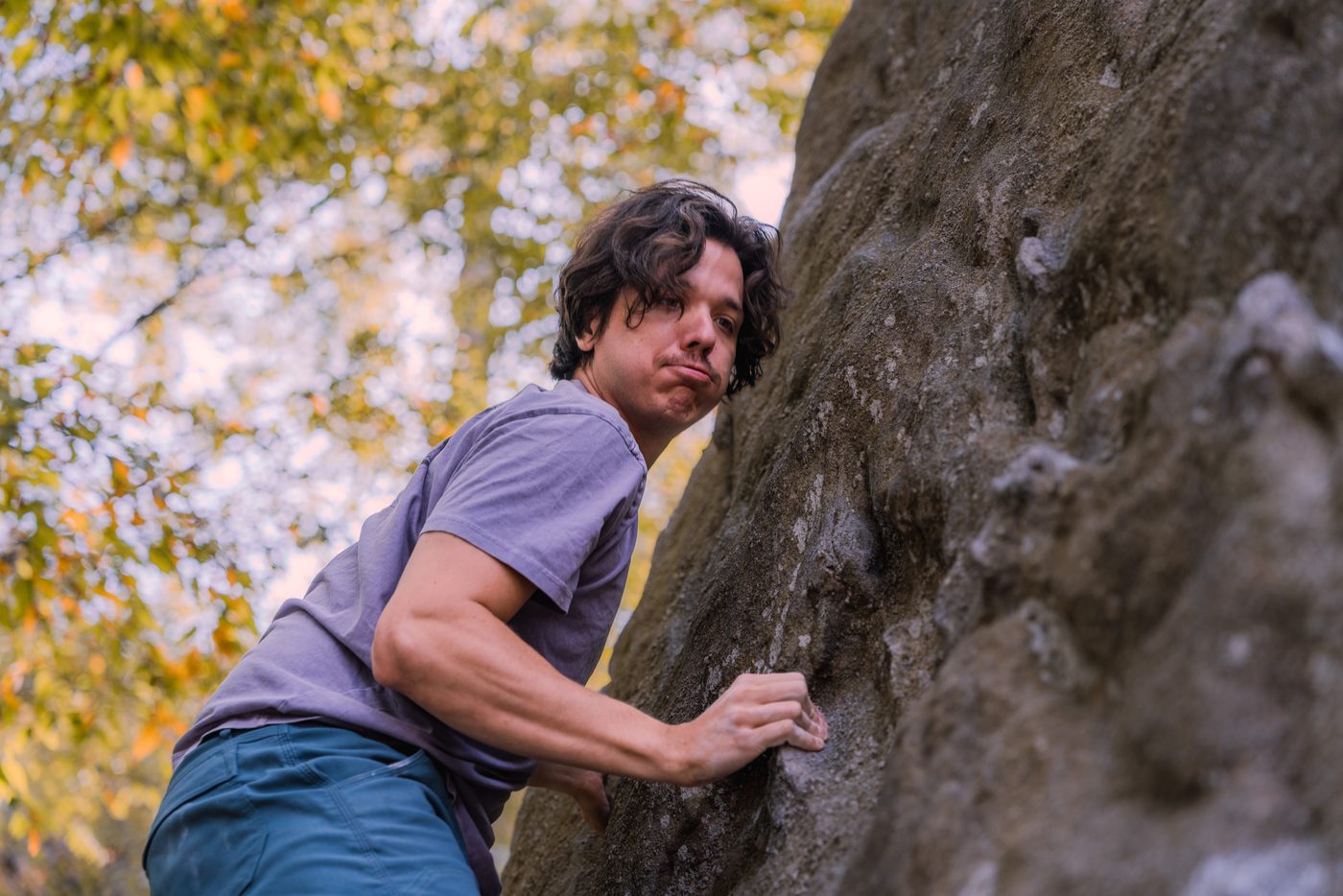A dark haired man in a faded purple tee crimps a hold on a lichen mottled boulder, lips pursed, backlit autumn leaves behind him.