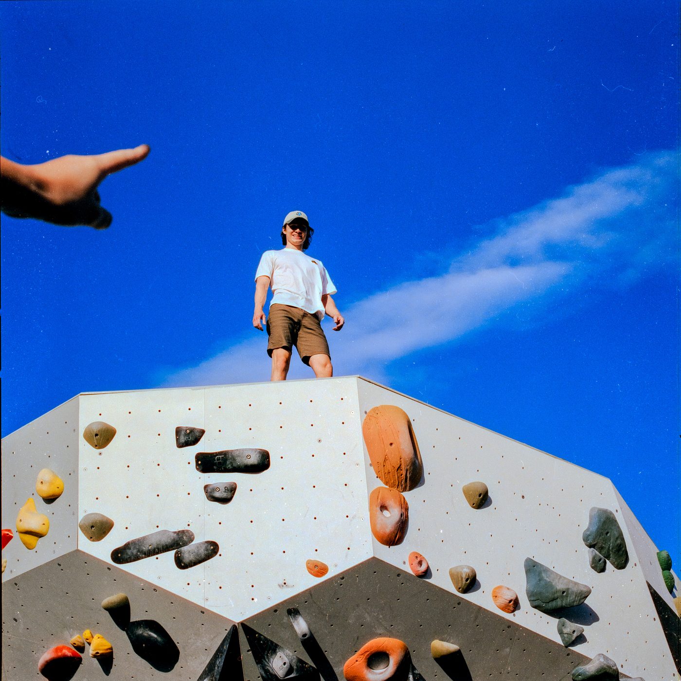 A man in a cap and white tee stands atop a hold studded bouldering wall against deep blue sky, a blurred hand pointing in from the corner.