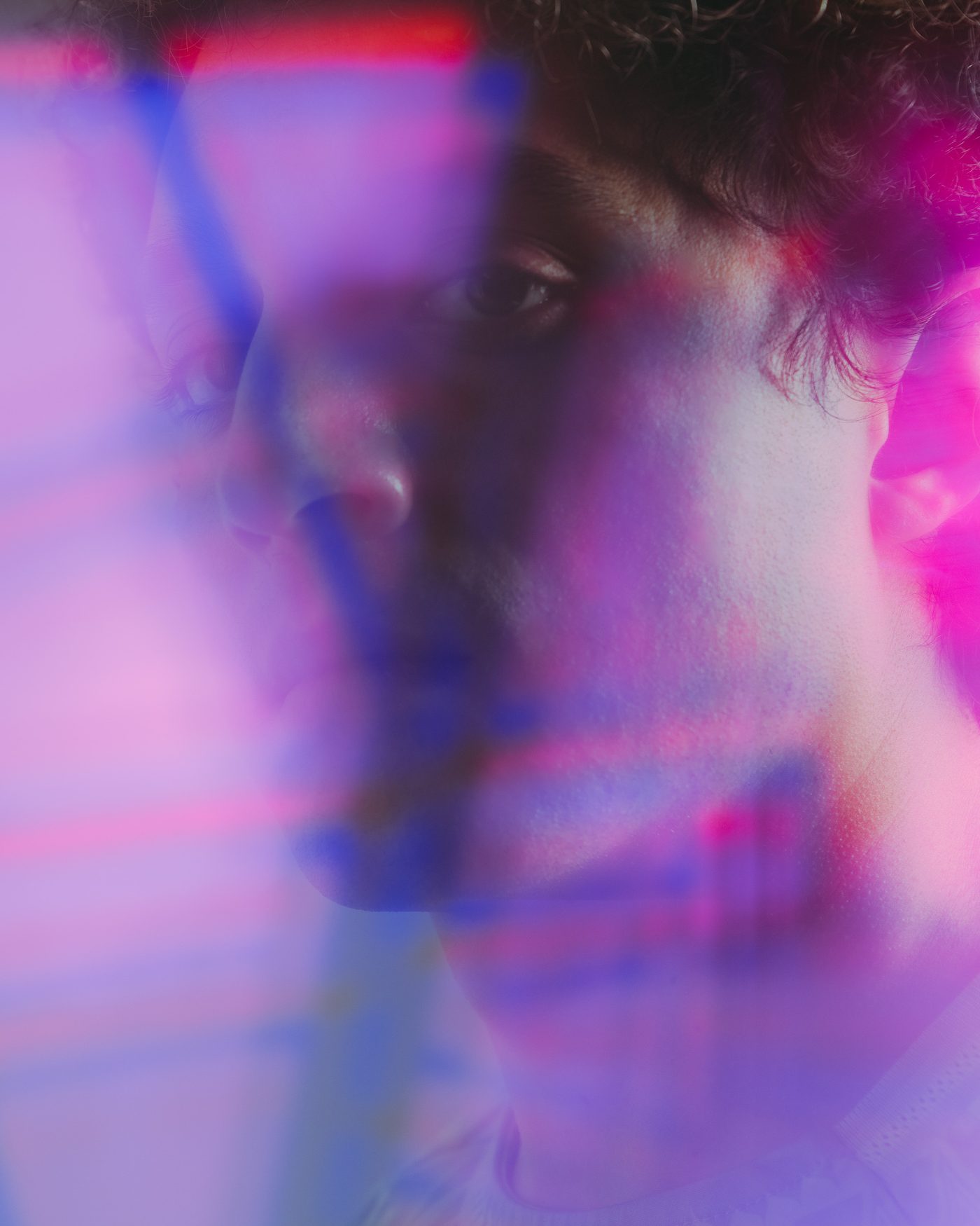 The photographer, a man with dark curly hair, turns his face away as magenta and violet prism light streaks across it, one eye still visible.