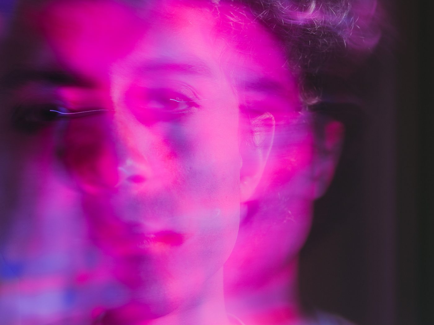 Lit in saturated magenta, the photographer's face fills the frame with eyes lowered, a doubled ghost of his profile offset to one side.