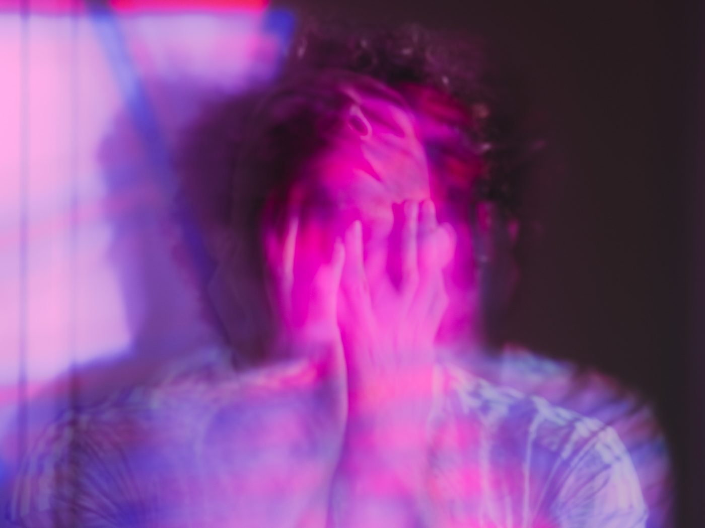 The photographer covers his face with both hands, fingers over his cheeks, his shirt and curls smeared into pink and violet blur.