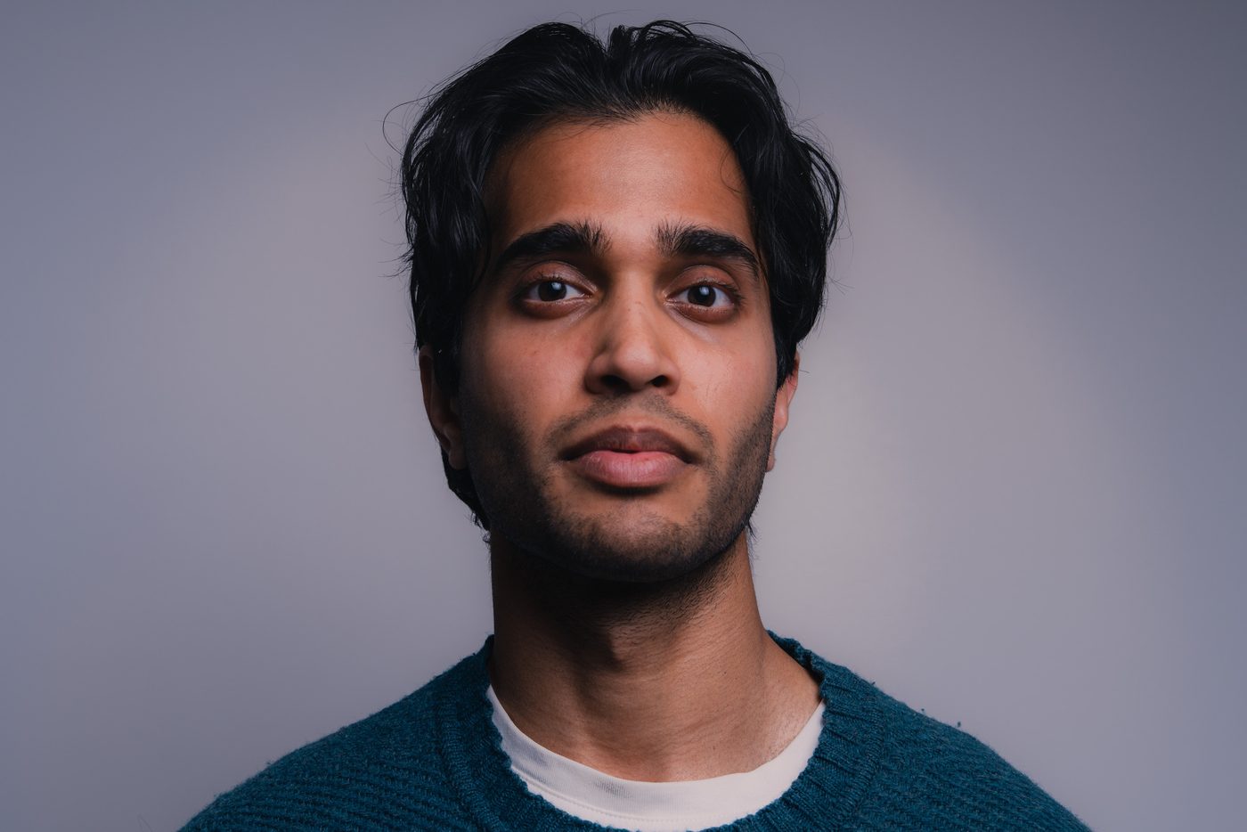 A man with thick dark hair and a teal knit sweater looks just past the camera, soft even light on his face against a grey backdrop.