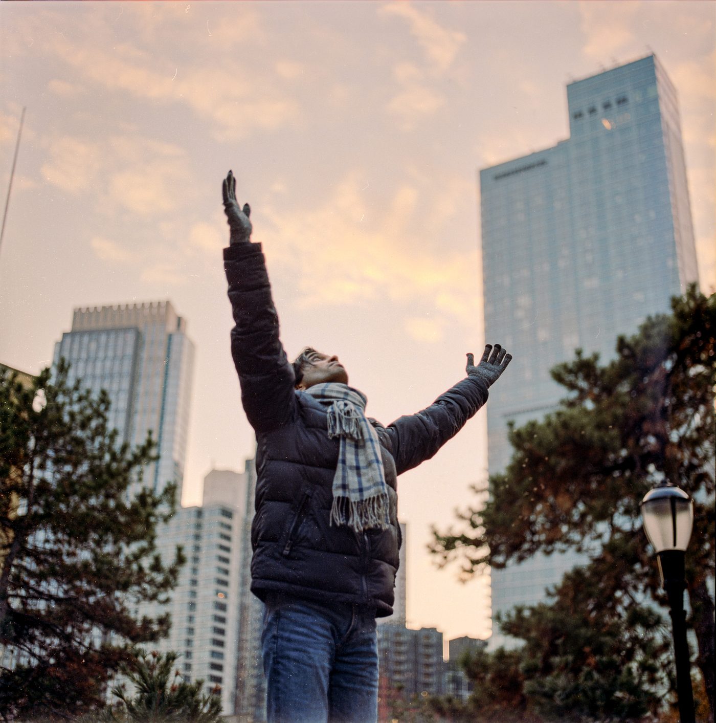 A man in a black puffer jacket and plaid scarf throws both arms wide and tips his head back beneath pink sunset clouds and city towers.